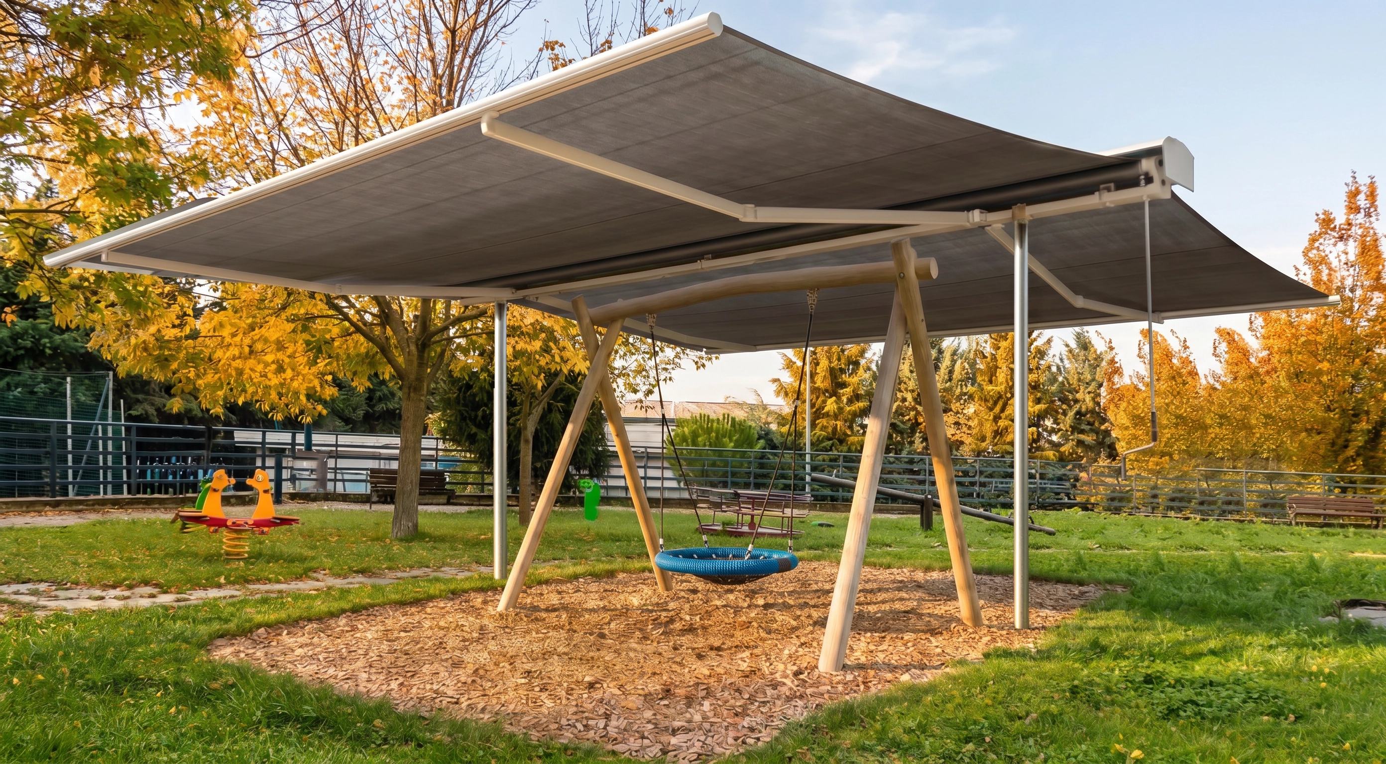 Ein moderner Spielplatz mit einer großen, grauen textilen Markise auf Holz- und Metallpfosten, die eine blaue Nestschaukel beschattet. Im Hintergrund Herbstbäume mit gelbem Laub, eine Wippe und ein Zaun bei sonnigem Wetter.
