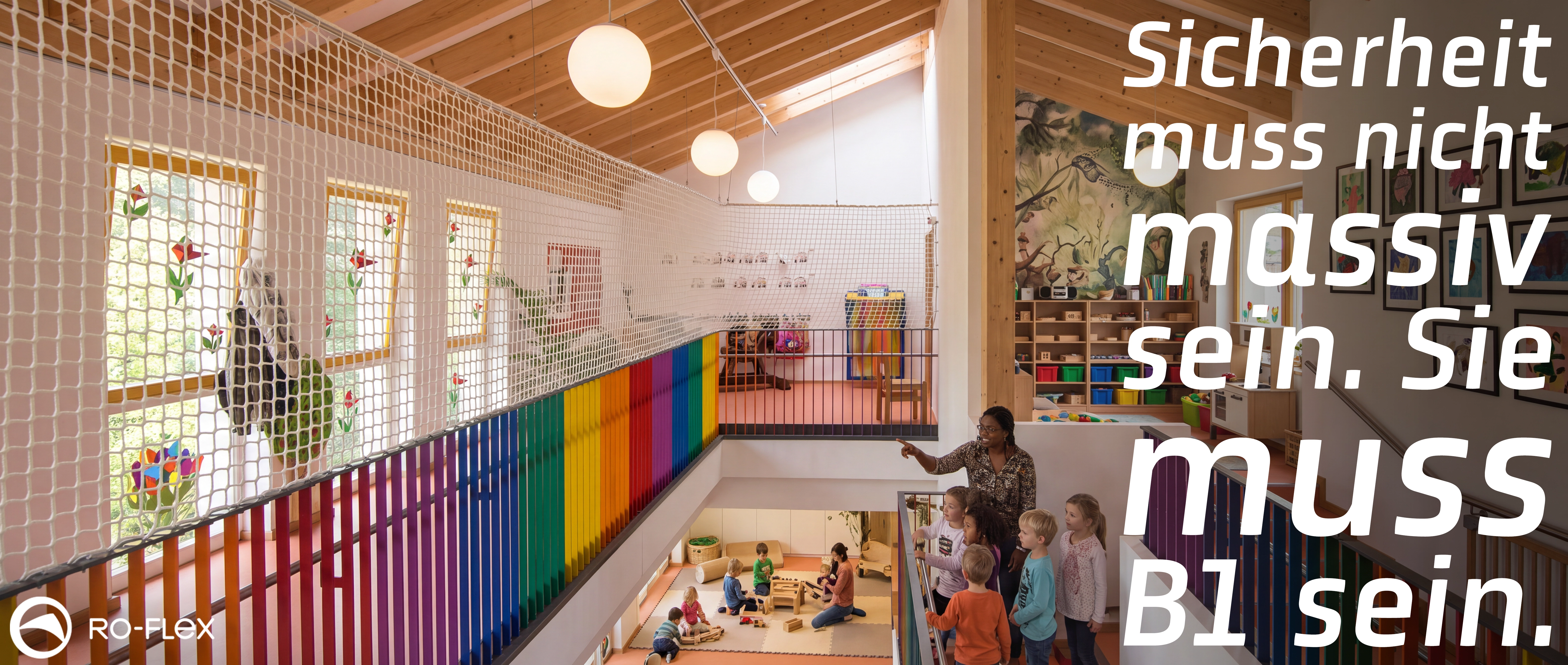 Helles, modernes Kindergarten-Atrium mit Galerieebene, gesichert durch weiße Sicherheitsnetze. Bunte Regenbogen-Brüstung, Kinder spielen im Erdgeschoss und stehen mit einer Erzieherin auf der Empore. Holzbalkendecke, große Fenster und RO-FLEX-Logo.